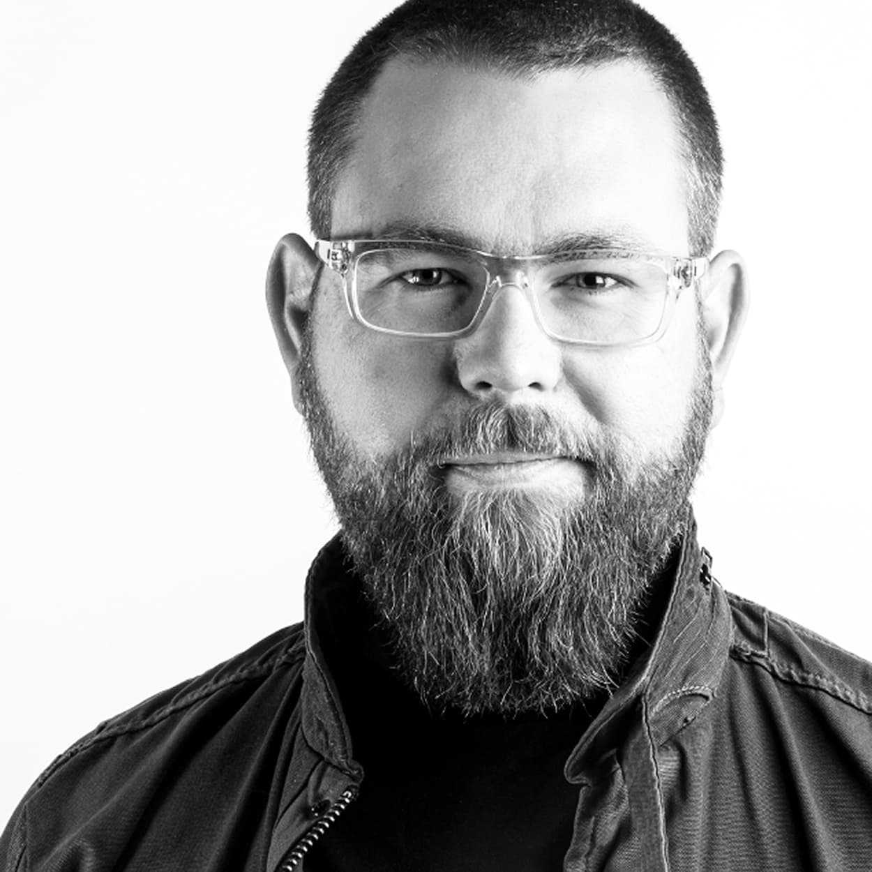 Man with a beard and clear-framed glasses, wearing a dark jacket, looking directly at the camera with a neutral expression.