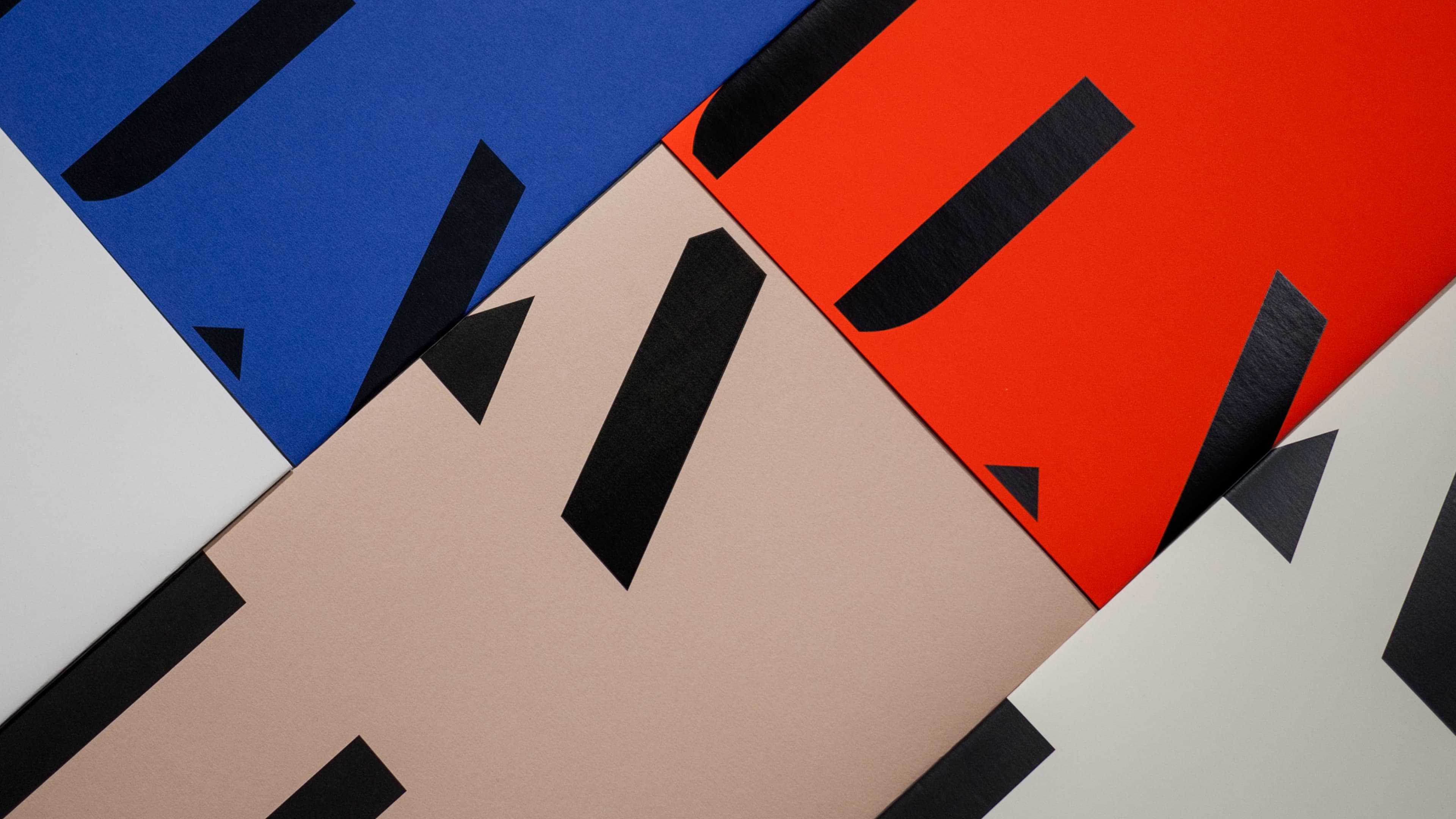 Close-up of overlapping printed cards or posters in blue, red, beige, and white, with large bold black letterforms and diagonal graphic elements.