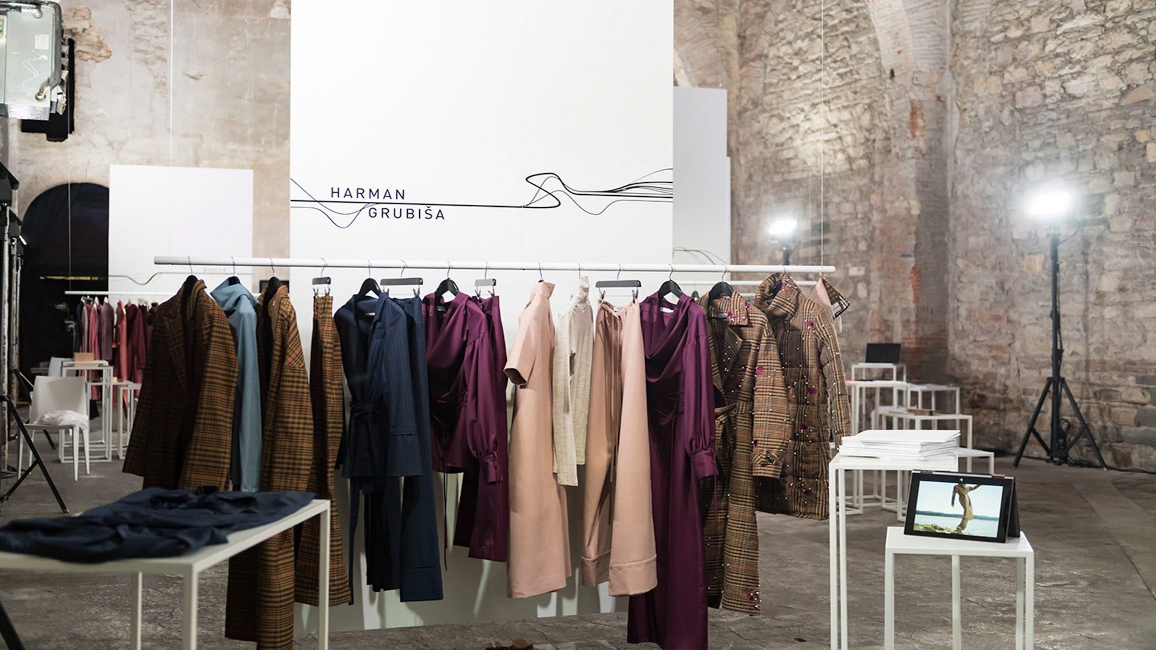 Harman Grubiša fashion presentation displaying coats, dresses, and outfits in burgundy, blush, navy, and plaid on a clothing rack, set within a stone-walled venue with white display tables and professional lighting.