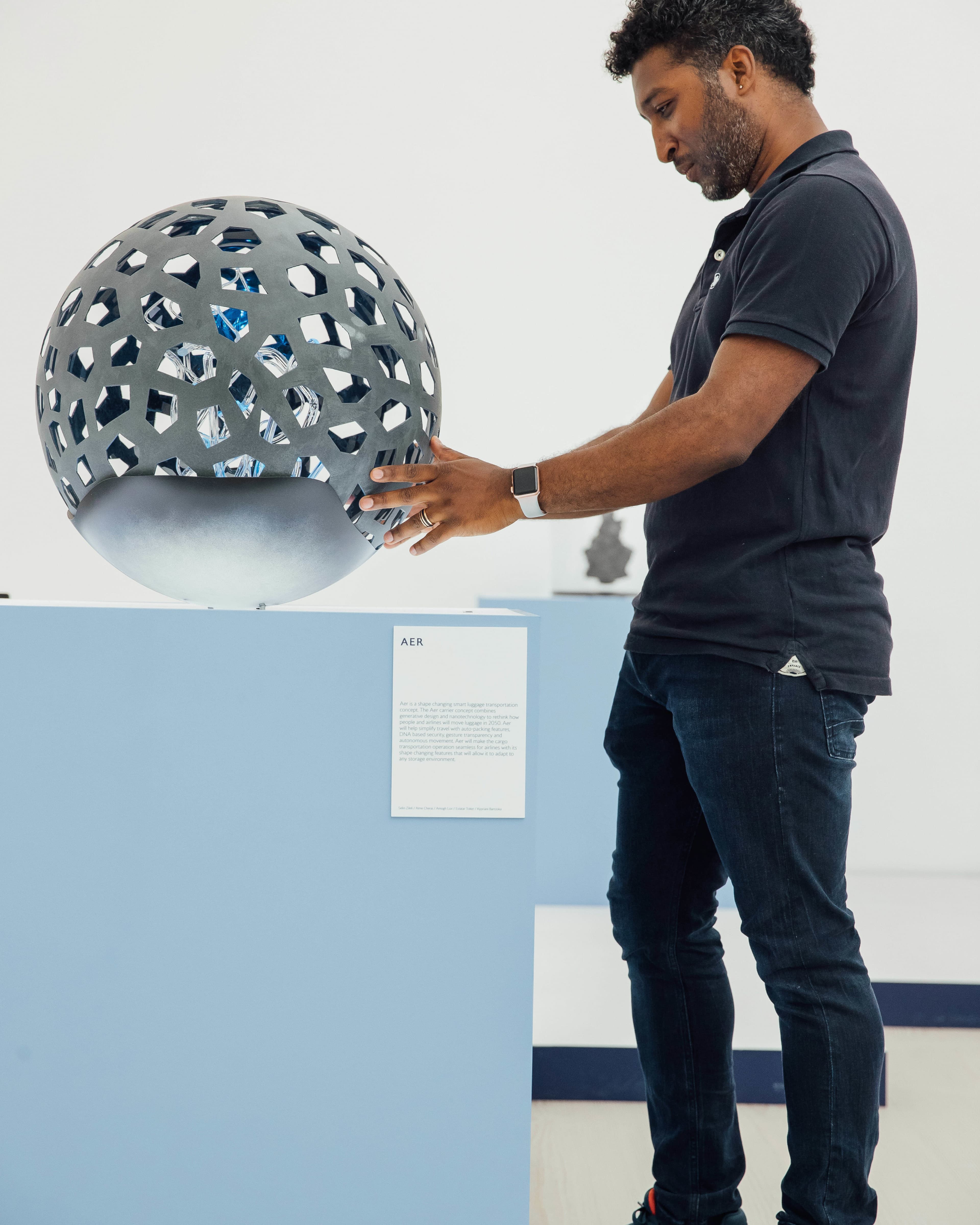 A man in a dark polo shirt and jeans examines a spherical sculpture with geometric cutouts revealing blue crystal-like interior, displayed on a light blue pedestal labeled "AER" in a gallery setting.