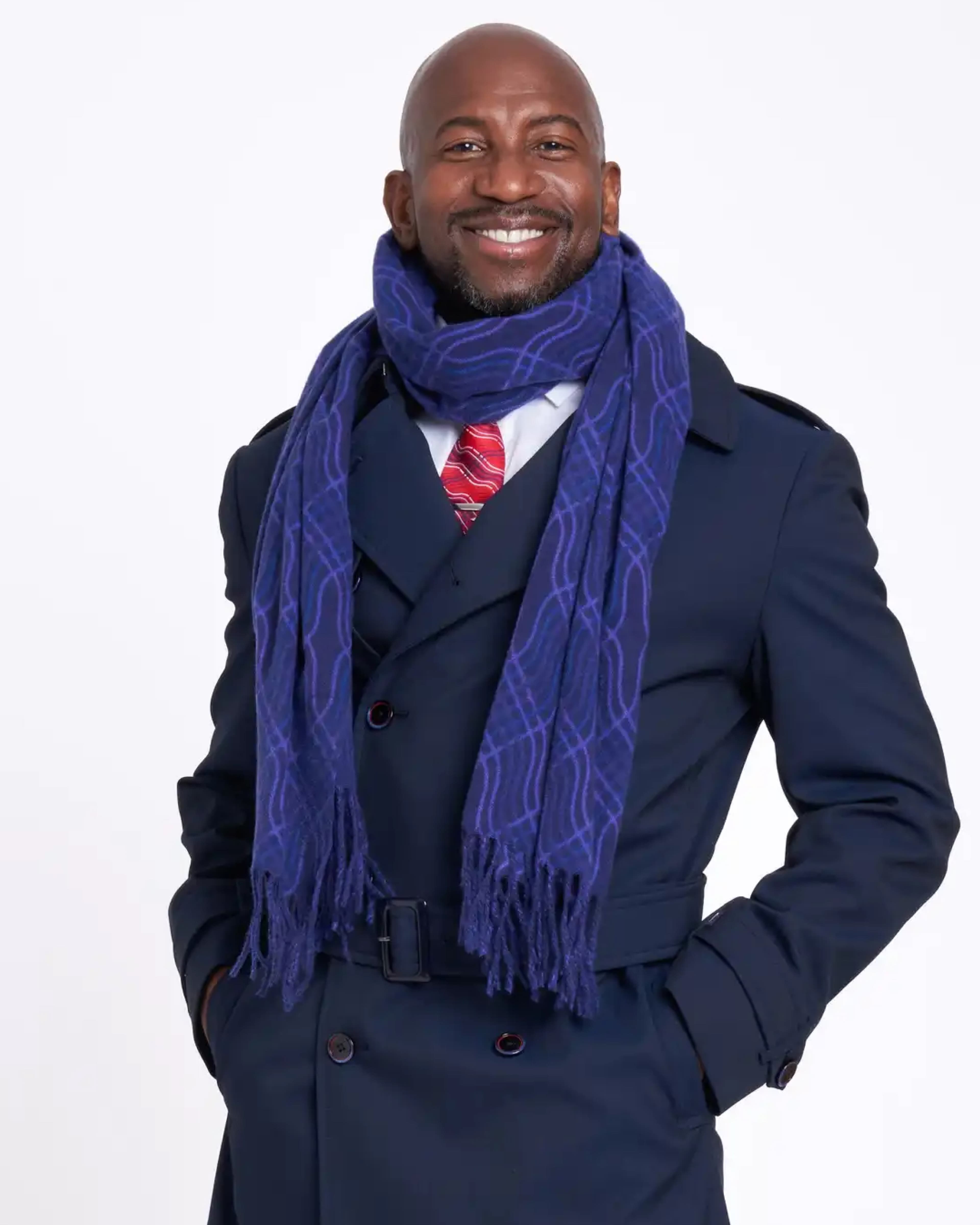 Smiling man in a navy double-breasted coat with a blue patterned scarf and red tie against a white background.