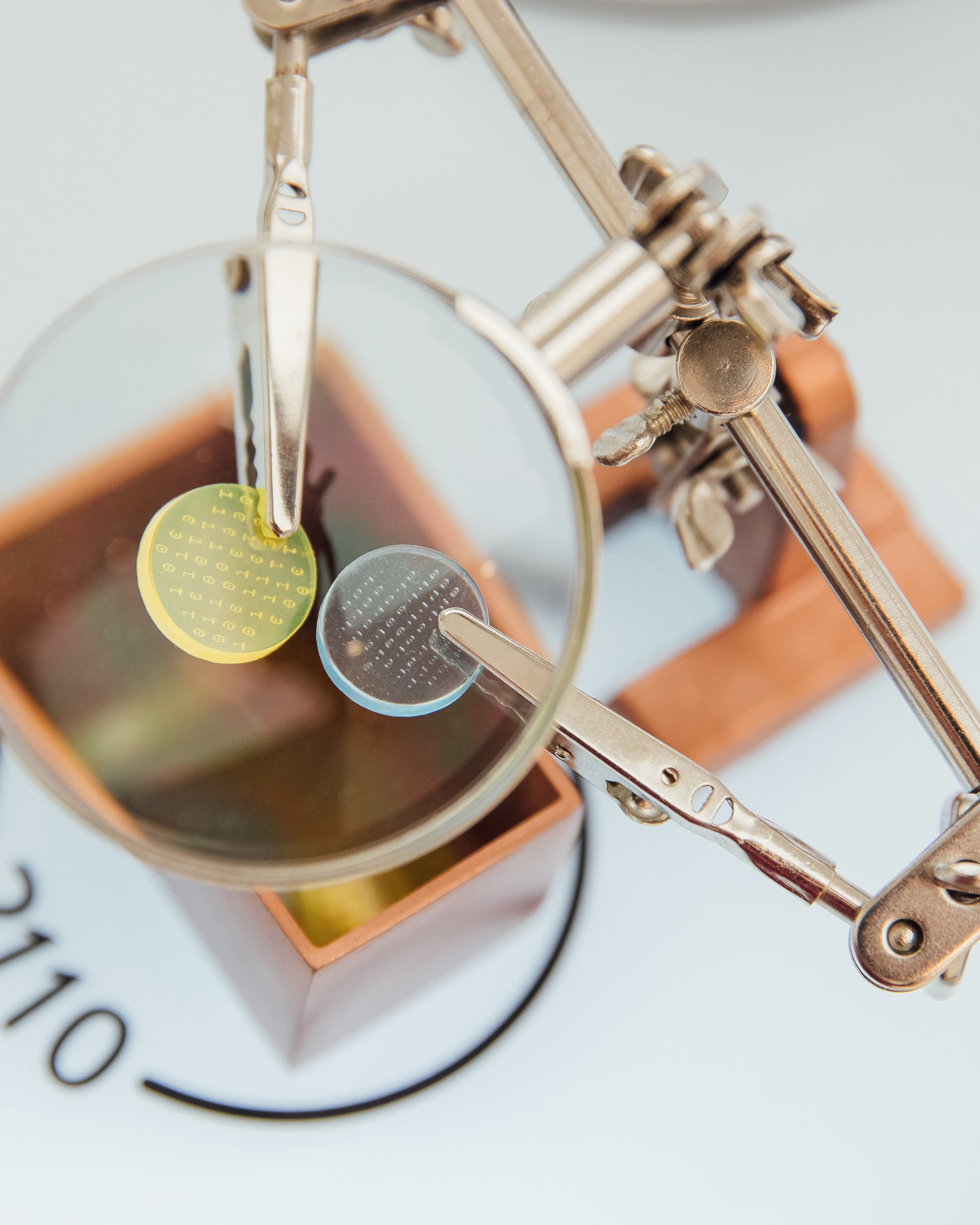 Two small circular chips — one yellow, one gray — held by crocodile clips on a helping-hands stand, viewed through a mounted magnifying glass from above.