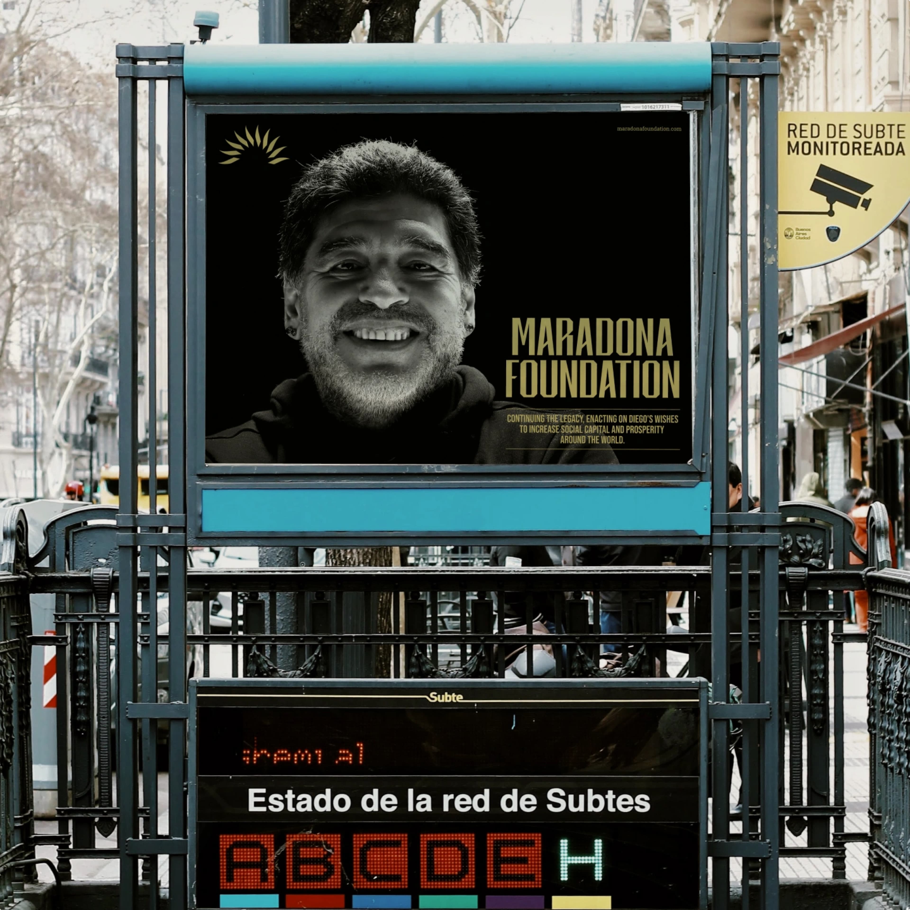 Billboard at a Buenos Aires Subte (subway) entrance featuring a smiling Diego Maradona promoting the Maradona Foundation, with the tagline about continuing his legacy to increase social capital worldwide.