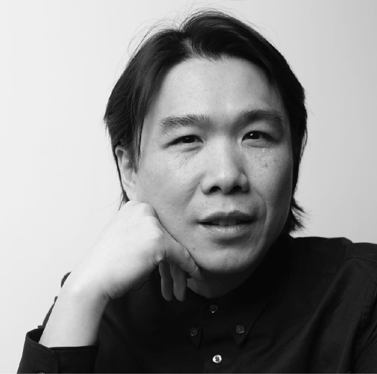 Black and white portrait of a person with chin resting on hand, wearing a dark shirt, looking at the camera.