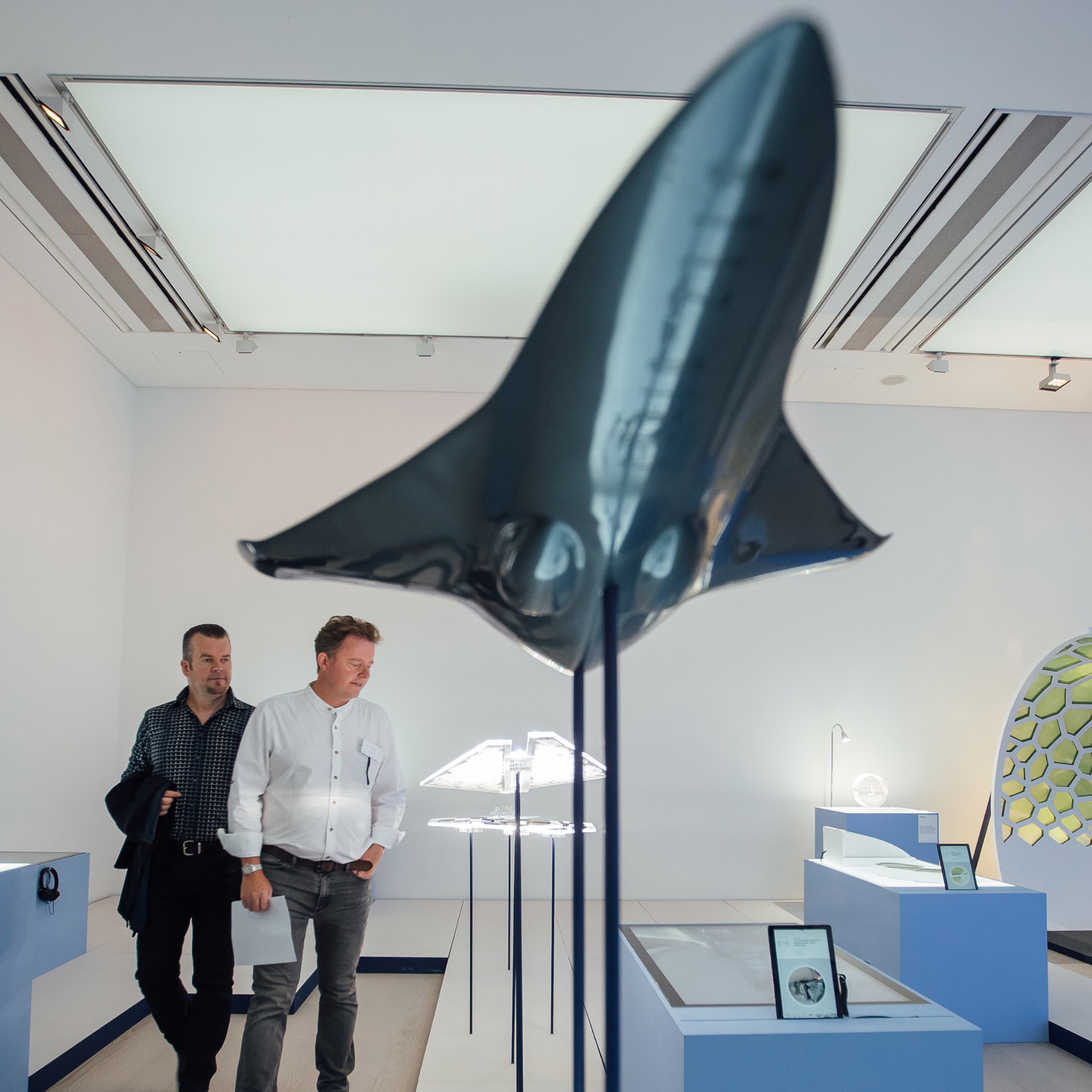Two men walking through a modern gallery space at the Saatchi Gallery, viewing exhibits including a large dark sculptural model of a plane or aircraft in the foreground and illuminated aviation-related displays on pedestals.