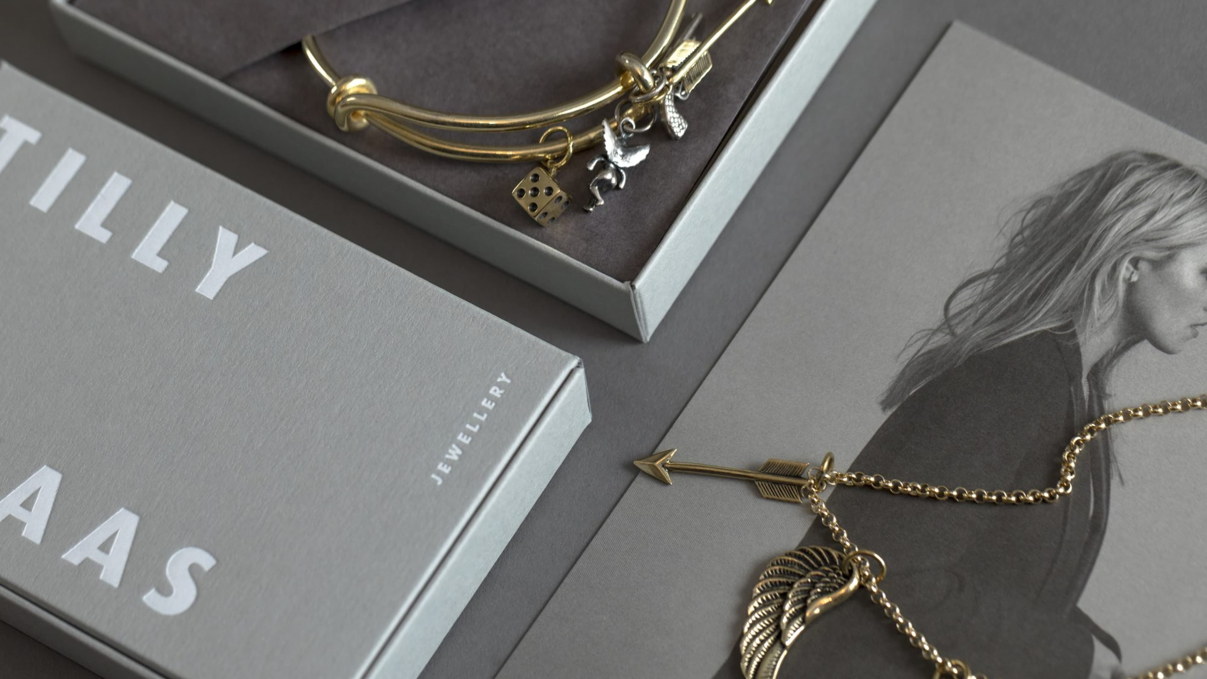 Tilly Haas jewellery: a gold bangle with silver charms including dice and a figure, displayed in a gift box, alongside a gold necklace with arrow and wing pendants, next to branded packaging and a monochrome brand image.