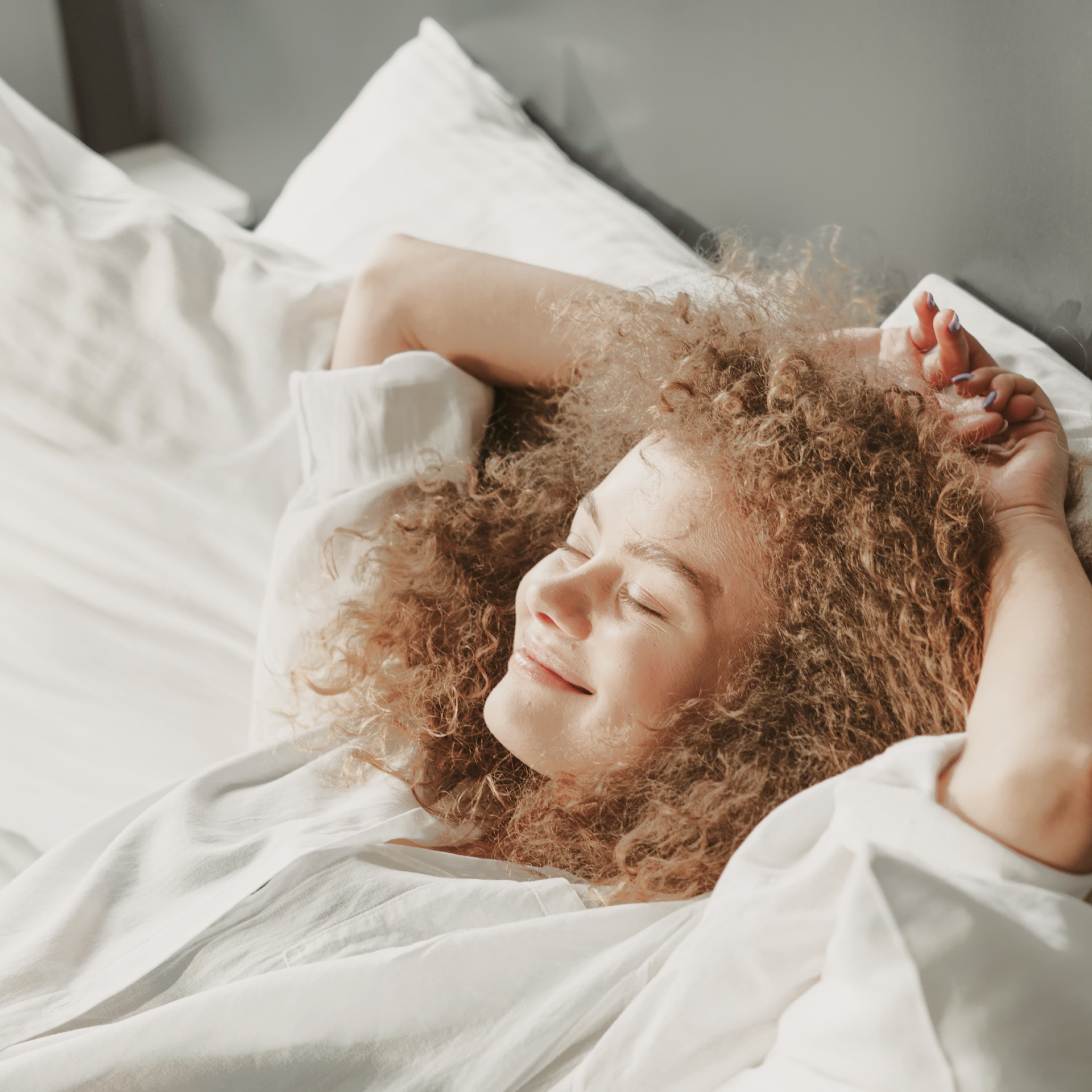 Person with curly hair smiling and lying on a white bed, wearing a white shirt, with arms behind their head, enjoying the morning light.