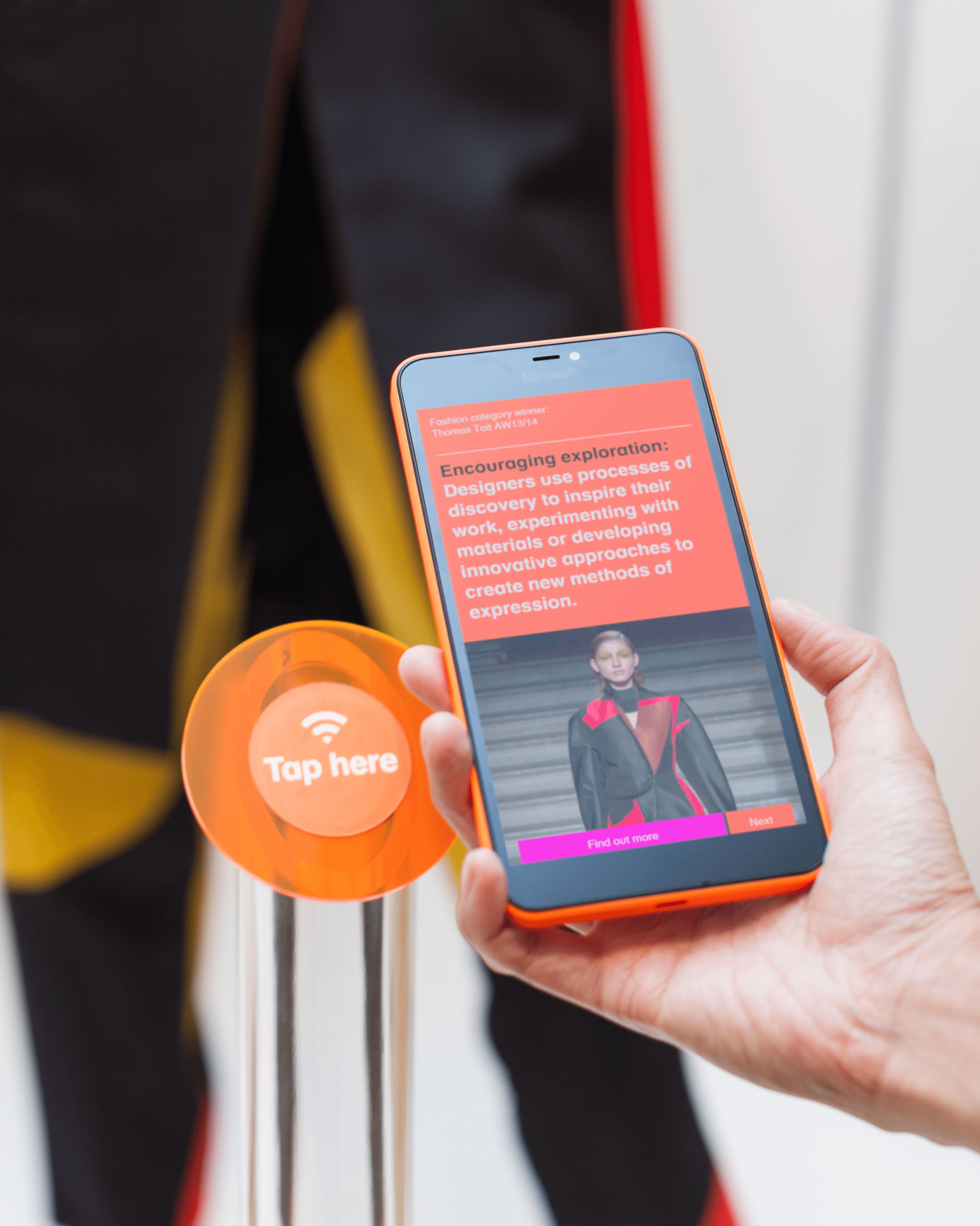 A hand tapping an orange NFC "Tap here" station with a smartphone, which displays an exhibition screen about the Fashion category winner Thomas Tait AW13/14, describing how designers use processes of discovery to create new methods of expression.