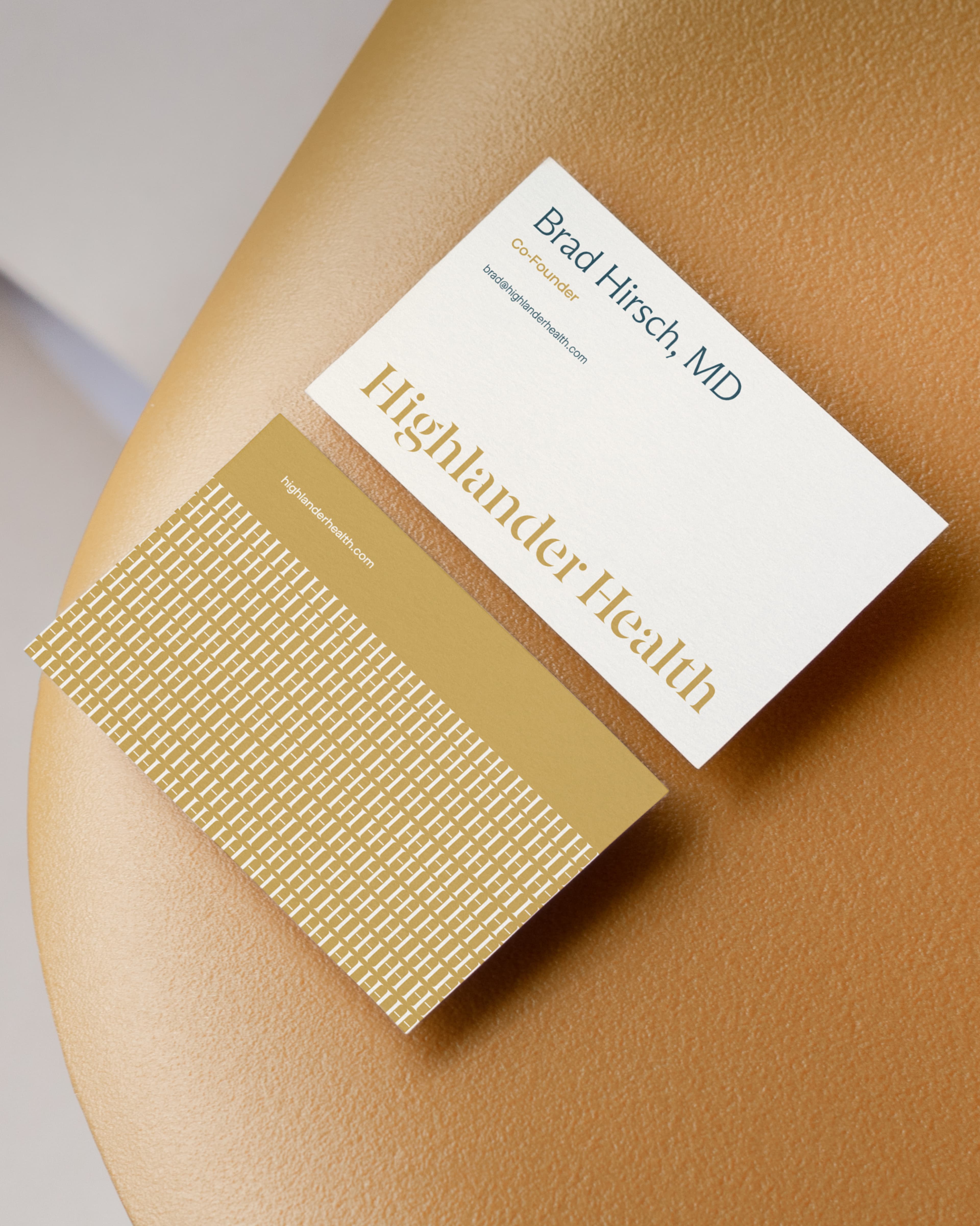 Two Highlander Health business cards on a golden surface: the front shows "Brad Hirsch, MD, Co-Founder" with email brad@highlanderhealth.com; the back features a gold geometric pattern with highlander health.com.