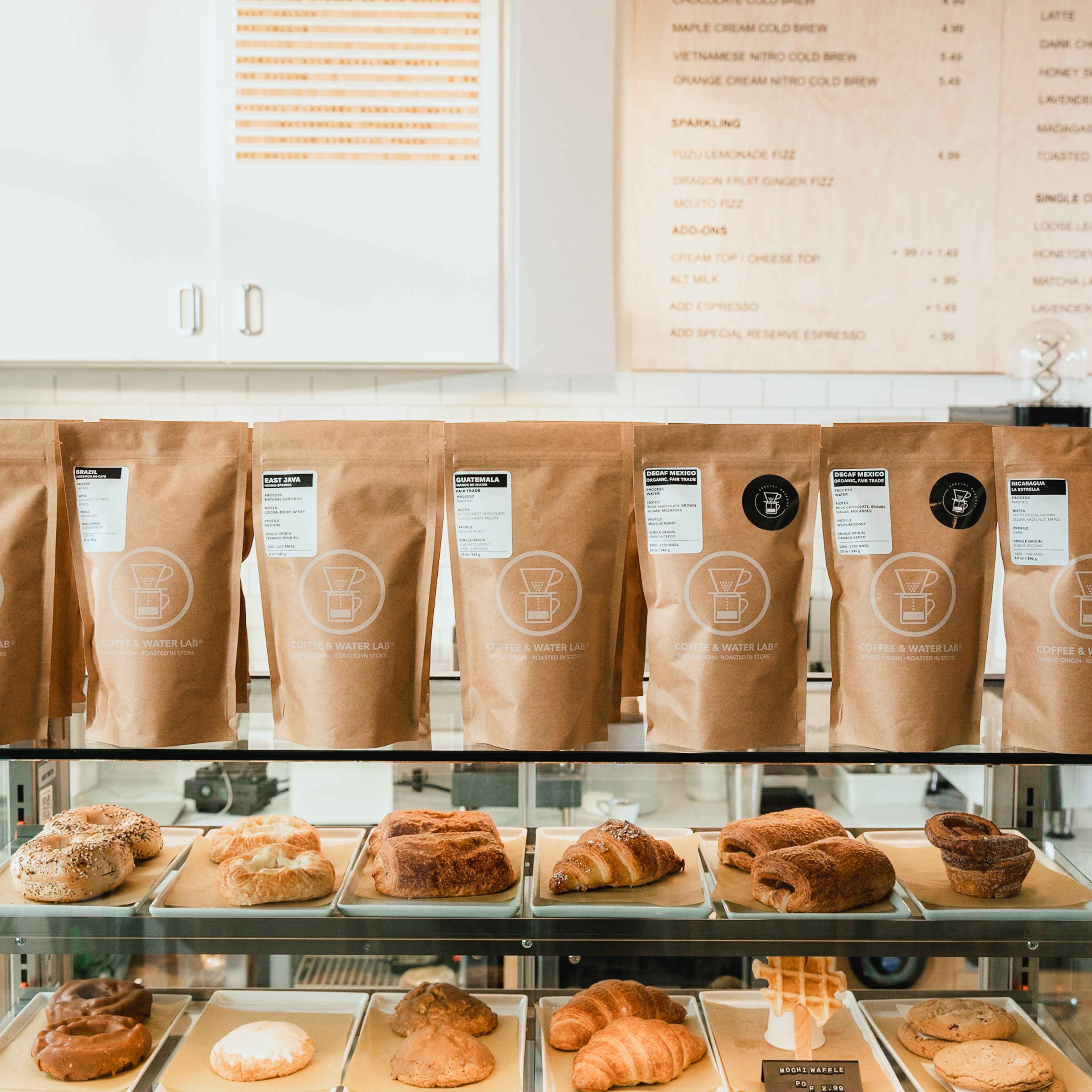 Coffee & Water Lab display counter with branded kraft paper bags of single-origin coffees including Brazil, East Java, Guatemala, Decaf Mexico, and Nicaragua, above a glass pastry case with croissants, bagels, pretzels, and other baked goods.