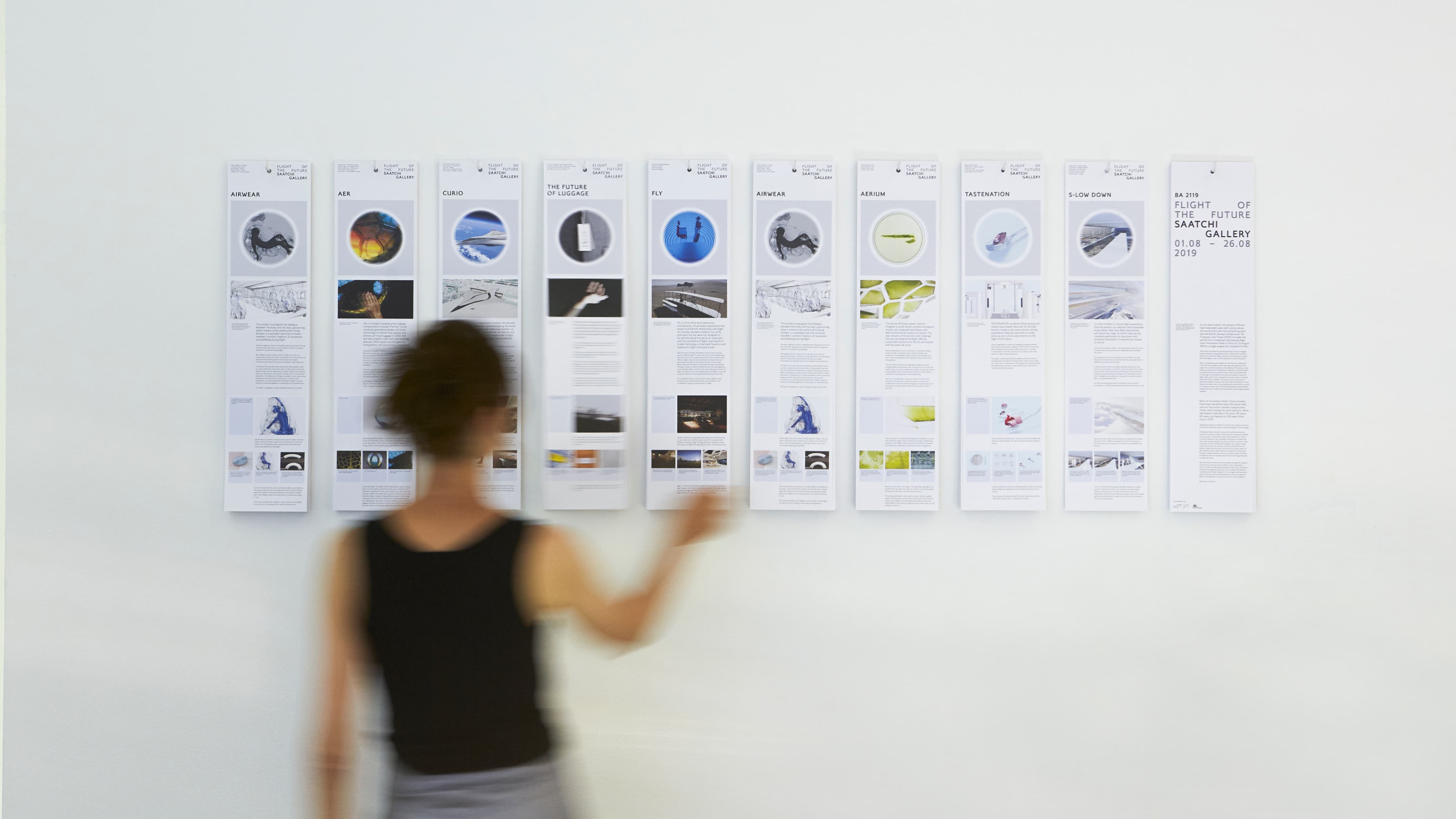 A visitor views a row of vertical project posters from the BA 2119 "Flight of the Future" exhibition at Saatchi Gallery (1–26 August 2019), featuring student concept designs including Airwear, Aer, Curio, The Future of Luggage, Fly, Aerium, Tastenation, and S-Low Down.