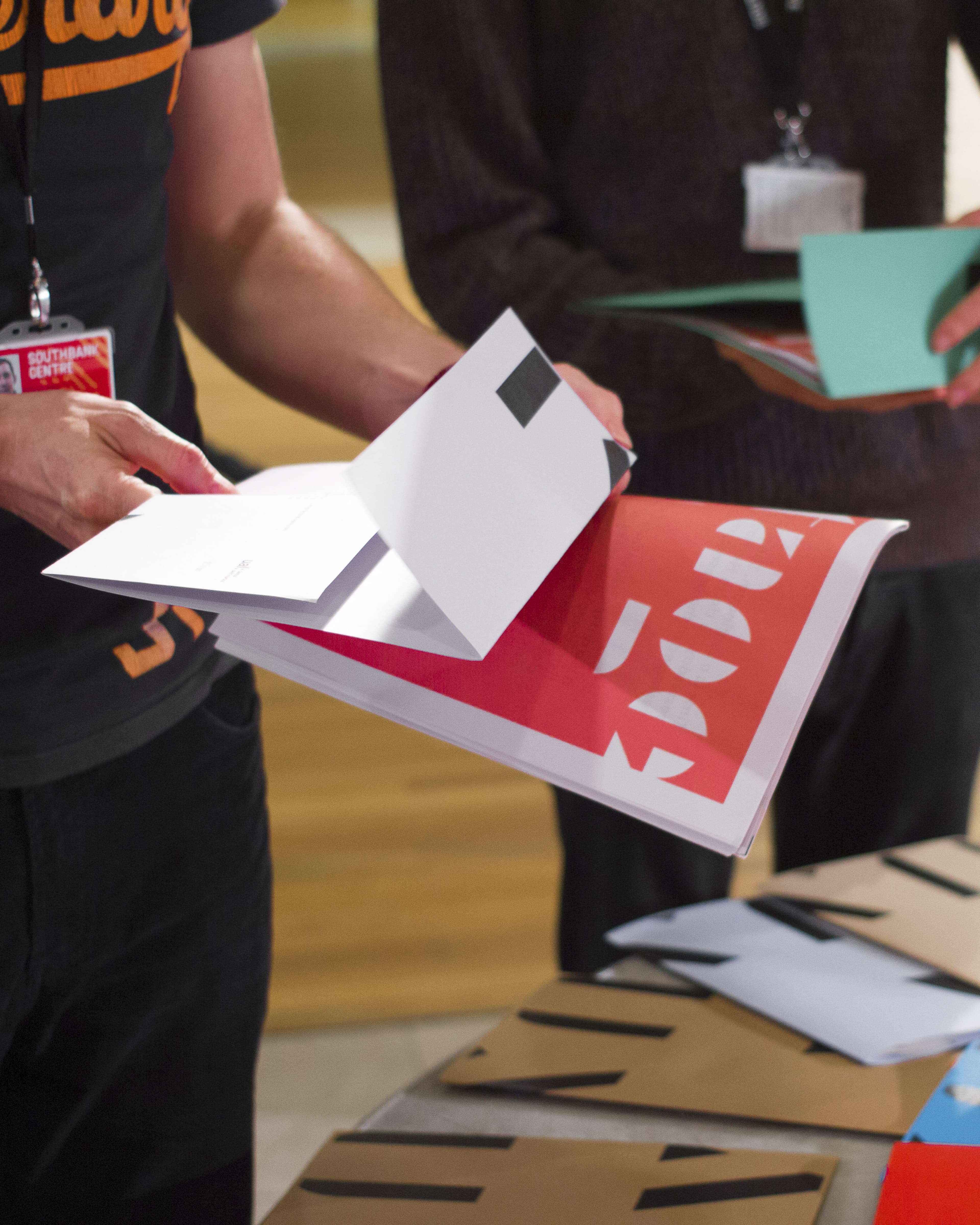 A person holding open a white booklet over a red graphic design brochure, wearing a Southbank Centre lanyard, at a table with printed materials.