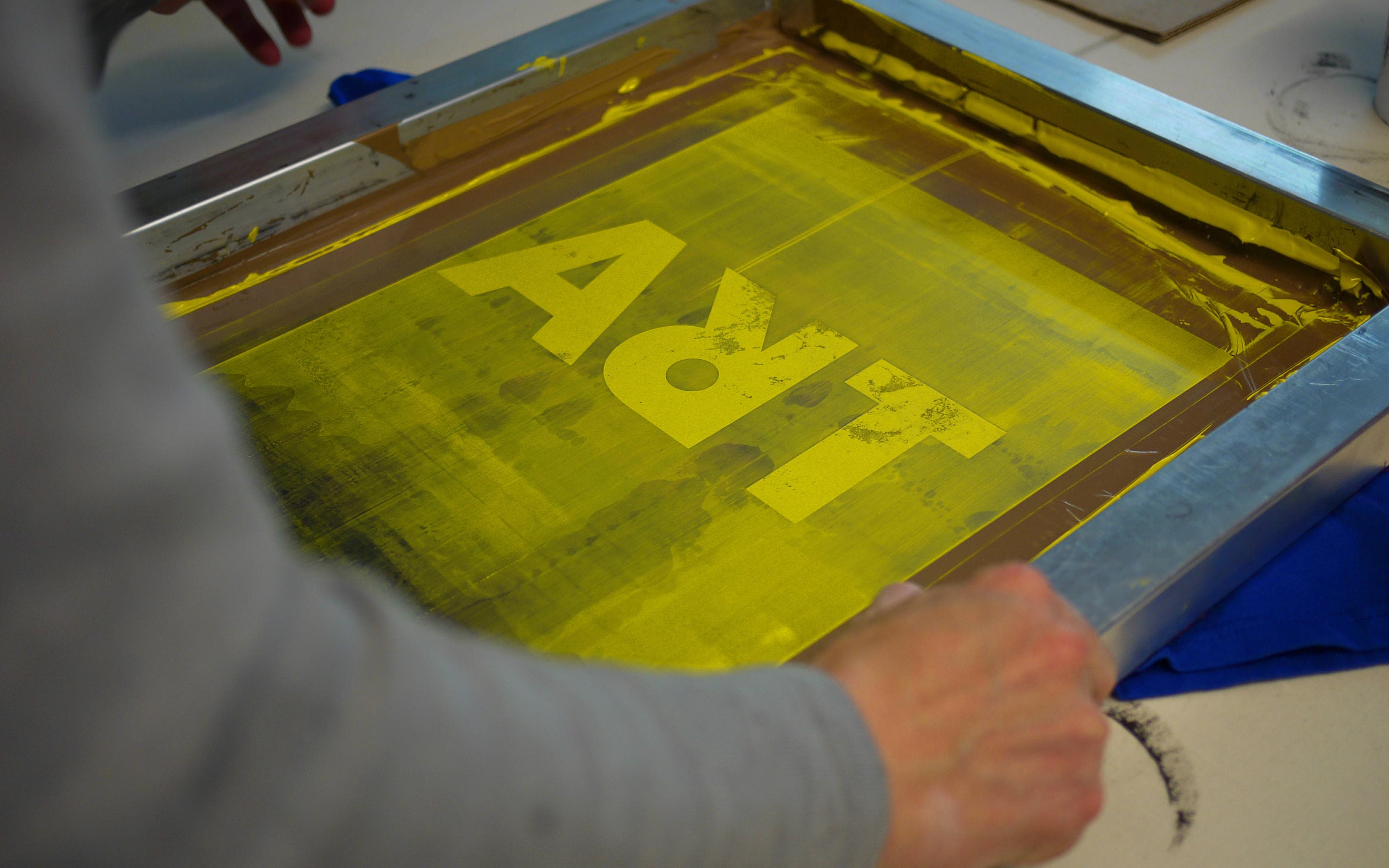 A person screen printing the word 'ART' using yellow ink on a screen in a studio setting.