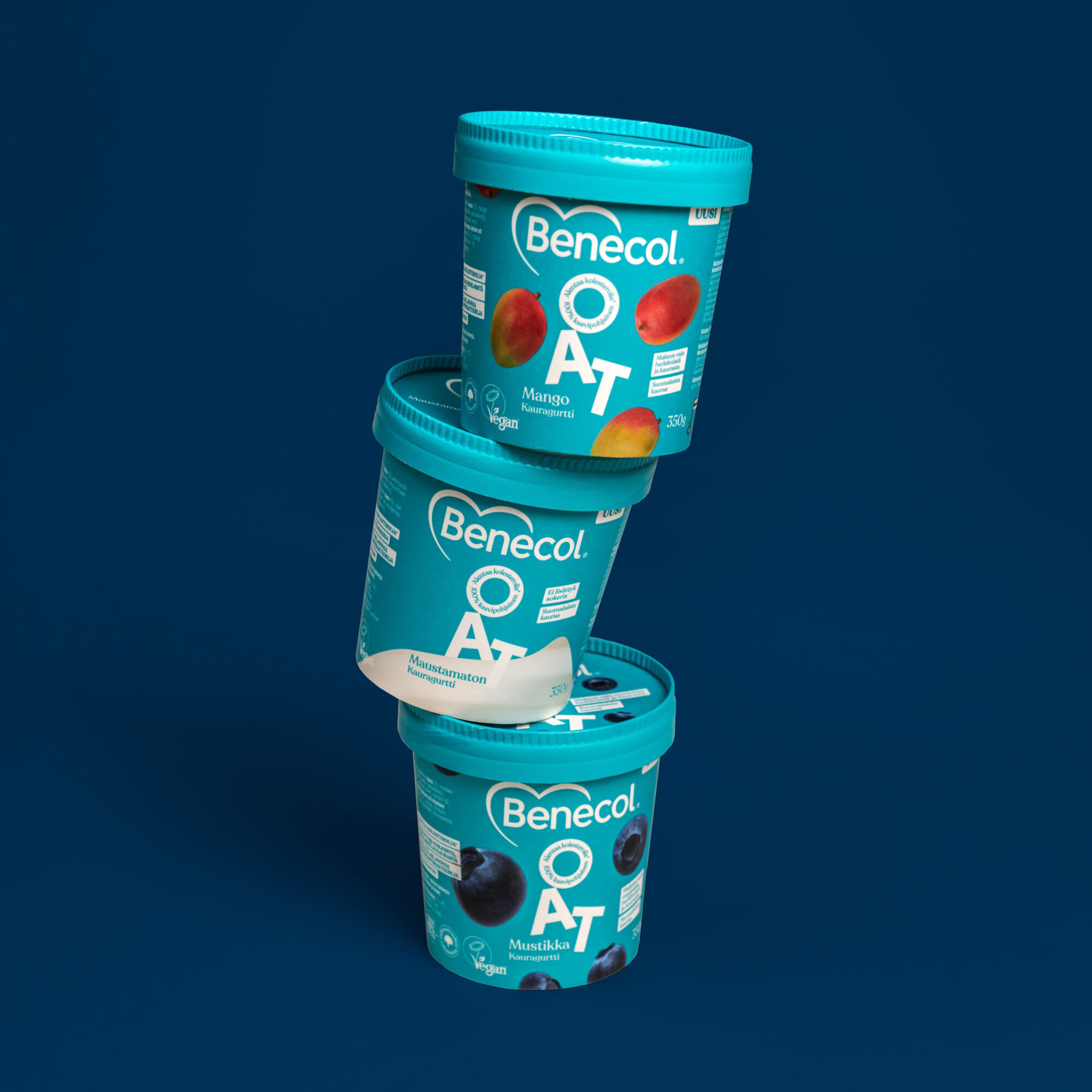 Three teal Benecol OAT Kauragurtti containers stacked on a dark blue background, showing Mango, Maustamaton (plain), and Mustikka (blueberry) flavors, all vegan.