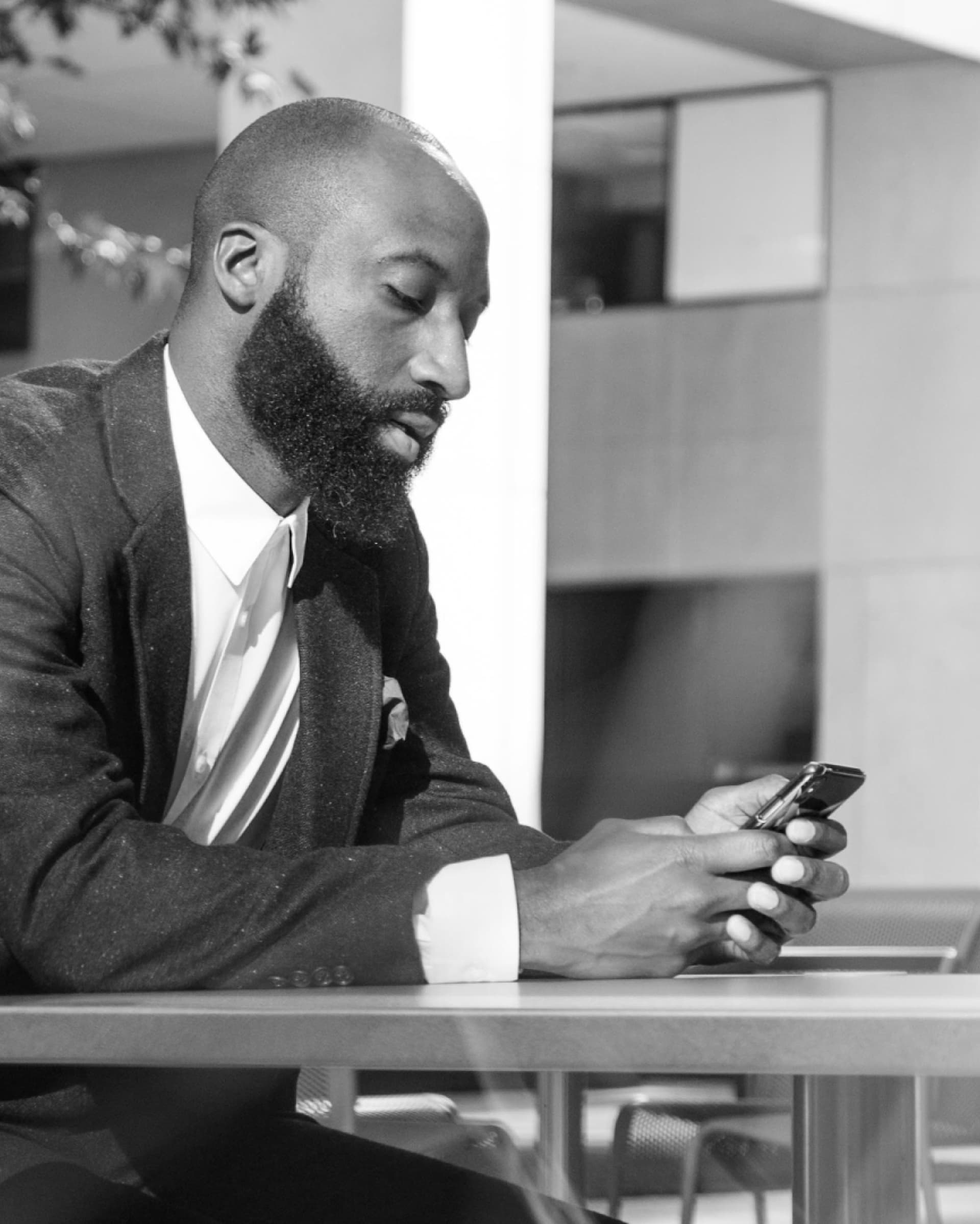 Black and white photo of a bearded man in a suit and tie sitting at an outdoor table, focused on his smartphone.