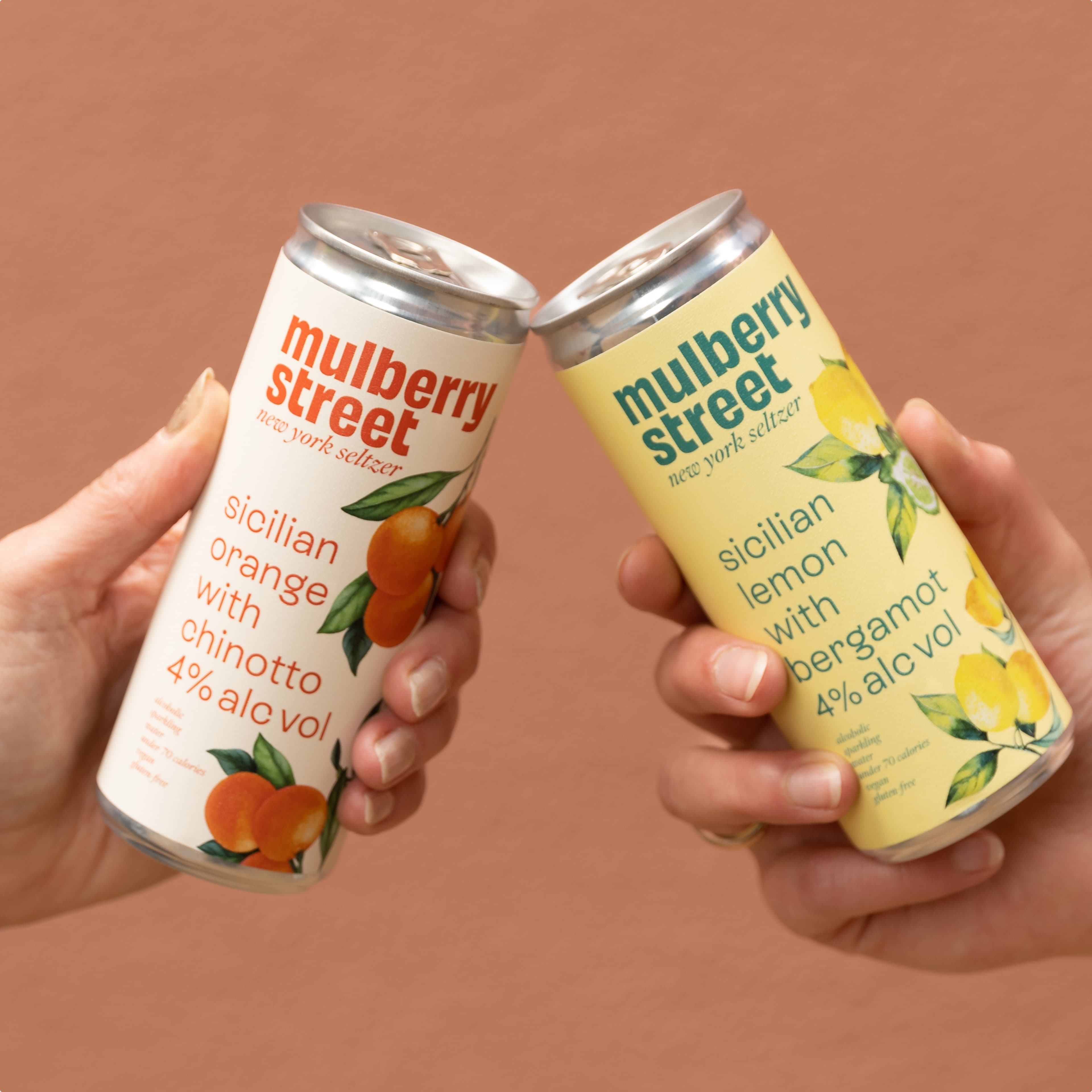 Two hands toasting with Mulberry Street New York Seltzer cans: Sicilian Orange with Chinotto and Sicilian Lemon with Bergamot, both 4% alc vol, on a warm terracotta background.