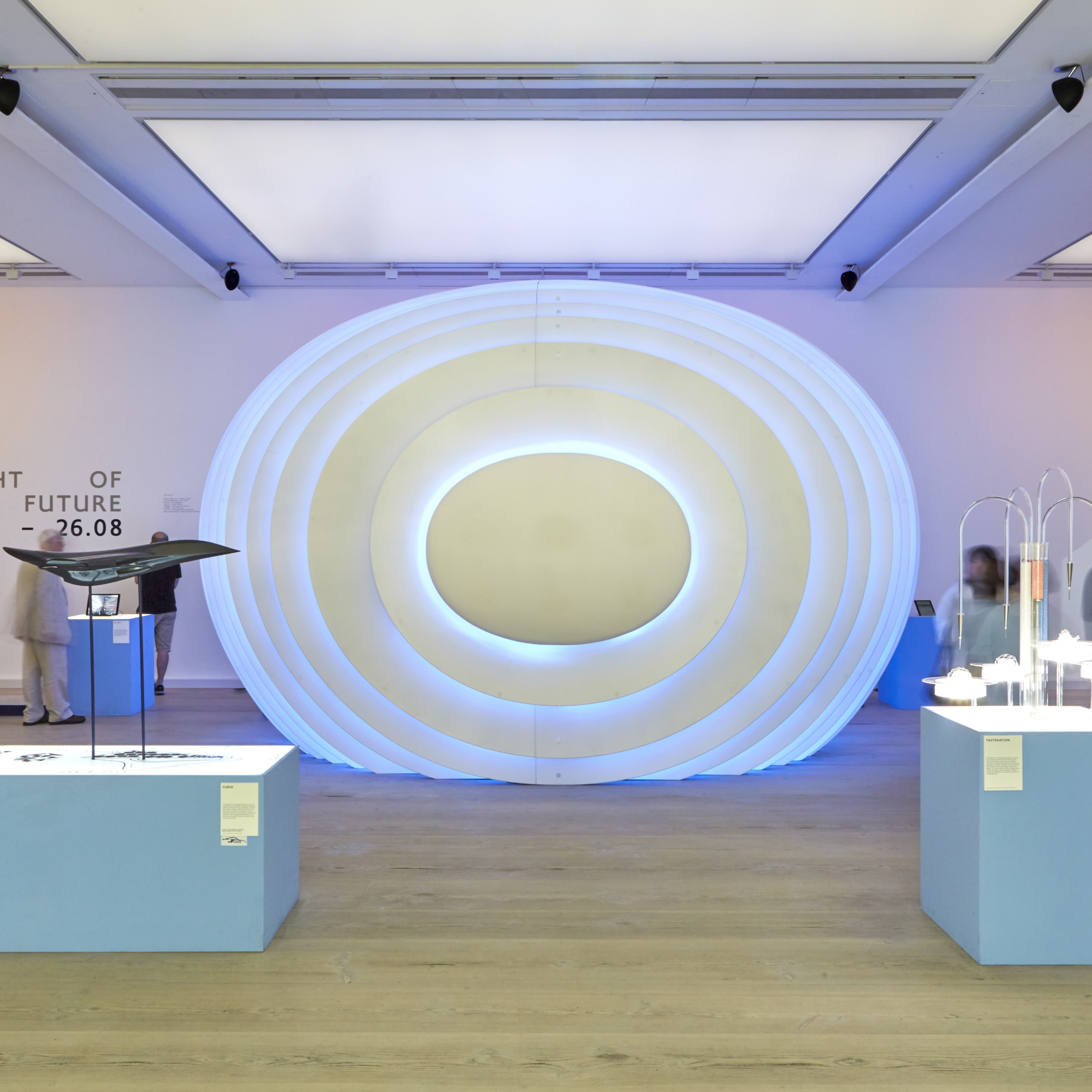 Museum gallery interior showing a large freestanding oval sculpture made of concentric white rings with blue LED lighting, flanked by light blue display pedestals with exhibit objects; partial text on wall reads "OF FUTURE – 26.08"